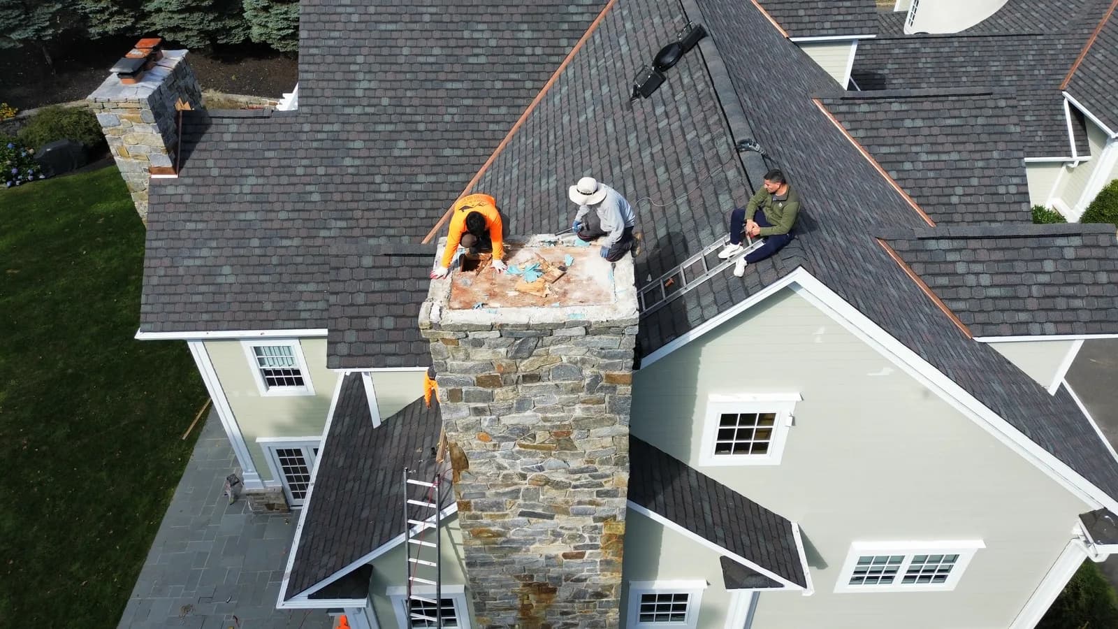 Stone chimney rebuild on a roof ridge with the crew working from a ladder in NJ